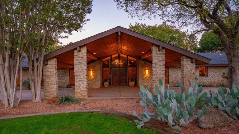 Stone house with a prominent wooden entrance, large pillars, and warm exterior lighting. Surrounded by trees, prickly pear cactus, and landscaped gravel, with a green lawn in the foreground.