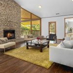 A spacious living room with stone walls, a lit fireplace, large windows with a view of greenery, light-colored sofas, a coffee table, yellow rug, and wooden floors. Natural light fills the room.