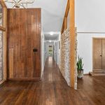 Spacious hallway in a modern home with wooden floors, wood-paneled and stone walls, double front doors, houseplants, and a glimpse into a bright living room.