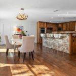 Bright, open kitchen and dining area with wooden floors, rustic wood cabinets, stone breakfast bar with clear stools, antler chandelier, round dining table with beige chairs, and large windows letting in natural light.
