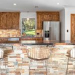 A rustic kitchen with wooden cabinets, a stone backsplash, and a matching stone island. Three metal wire barstools sit at the island, and stainless steel appliances are visible. A window overlooks a green outdoor area.