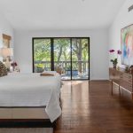 A spacious bedroom with wood flooring, a large bed with white bedding and leopard print pillows, a dresser with flowers, a large mirror, colorful wall art, and sliding glass doors that open to a patio with trees outside.