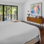 A bright bedroom with a large bed, white bedding, leopard print pillows, a wooden dresser with plants and decor, a colorful abstract painting, a large mirror, and glass doors opening to a balcony with trees outside.