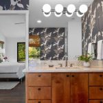 A bathroom with bird-patterned wallpaper, a wooden vanity with a marble countertop, a plant, two mirrors, and globe lights above. An open door reveals a bedroom with a bed and window.