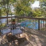 A wooden deck with a round glass table and four chairs overlooks a backyard pool and a lake surrounded by trees on a sunny day. Potted plants decorate the deck railing and table.