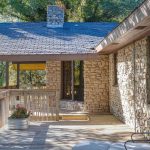 A stone house with a wooden deck features outdoor seating and potted flowers. Trees provide shade, and sunlight filters through, creating a cozy, inviting atmosphere.