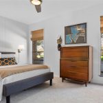 A cozy bedroom with a single bed, white bedding, a tan throw blanket, a geometric pillow, a tall wooden dresser, a guitar by the window, and large windows showcasing trees outside. Art and hats hang on the wall.