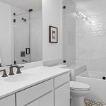 Modern white bathroom with a glass-enclosed bathtub and shower, white vanity with dark fixtures, green soap dispenser, potted plant, towels on the wall, and geometric tile floor.