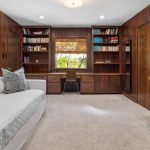 A cozy home office with wood-paneled walls, built-in bookshelves, a built-in desk under a window with a chair, a white sofa with pillows, carpeted floor, and a wall-mounted TV.