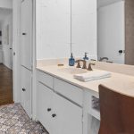 A bathroom with a white vanity, beige countertop, and a wooden chair. A tray with colorful flowers and soap dispenser sit on the counter. Patterned tile floor and a towel are also visible. A hallway is seen through the open door.