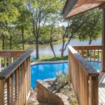 A wooden deck with railings overlooks a clear blue swimming pool, lush green grass, trees, and a calm lake in the background under a sunny sky.