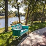 A turquoise bench sits on grass beside a swimming pool, overlooking a calm river bordered by trees and greenery under a clear sky.