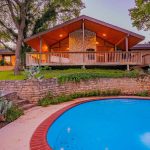 A stone house with a wrap-around wooden porch is surrounded by trees and warm lights at dusk. A curved swimming pool with red brick trim sits in the foreground, bordered by stone landscaping and cacti.