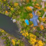 Aerial view of a house with a blue swimming pool surrounded by autumn trees near a riverbank, with a small dock on the water and a red-roofed building nearby.