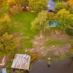 Aerial view of a house surrounded by trees, a grassy yard, a small swimming pool, and a wooden dock extending over the edge of a pond or lake at sunset.