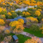 Aerial view of a house surrounded by trees with autumn foliage, a winding driveway, and a small pond or lake in the background, under a clear sky.