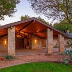 A stone house with a wide wooden A-frame roof, lit porch, and large windows, surrounded by trees, cacti, and a neatly landscaped yard at dusk.
