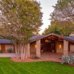 A single-story stone house with a covered entryway, surrounded by mature trees, a curved driveway, and desert landscaping with cacti and shrubs at sunset.