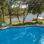A backyard features a clear swimming pool, surrounded by grass and trees, with a view of a calm river and a small wooden dock in the background under a sunny sky.
