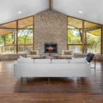A spacious living room with stone walls, a vaulted ceiling, large windows, a central fireplace, a white sofa, armchairs, and a wooden floor, overlooking a green outdoor area.