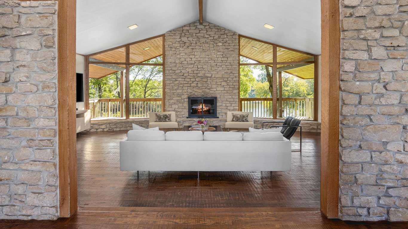 A spacious living room with stone walls, a vaulted ceiling, large windows, a central fireplace, a white sofa, armchairs, and a wooden floor, overlooking a green outdoor area.