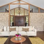 A modern living room with a white sofa, wooden coffee table, and yellow rug. The background features a stone accent wall, large double wooden doors, and a tall plant in a metallic pot. The ceiling has exposed wood beams.