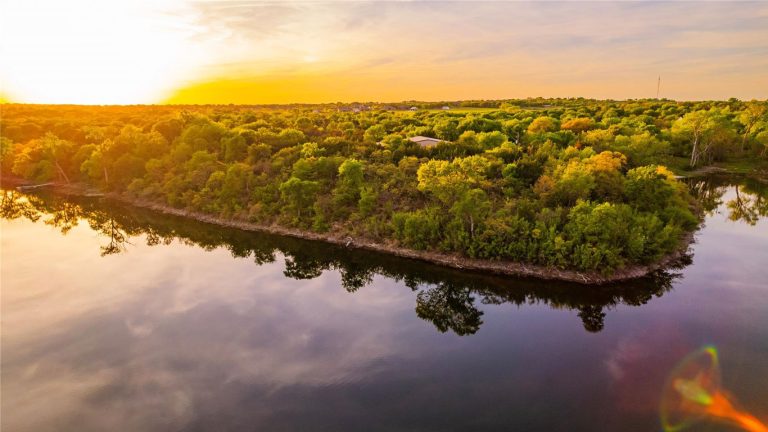 A golden sunset reflects off calm water surrounding a lush, tree-covered peninsula. The sky glows with warm hues, and dense greenery stretches across the landscape under soft light.