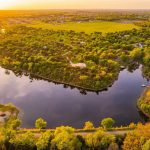 Aerial view of a serene lake surrounded by lush green trees and scattered houses at sunset, with fields and a small town visible in the background.