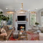A bright, modern living room with a brown sofa, white armchair, round dining table with white chairs, fireplace with TV above, large windows, indoor plants, and a gold chandelier hanging from the ceiling.
