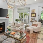Bright, eclectic living room with a lit fireplace, wall-mounted TV, white armchair, round dining table with white chairs, patterned rugs, decorative chandelier, and large windows letting in natural light.