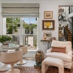 Bright, stylish living space with a round glass table, four white chairs, a plush armchair, and eclectic decor. Sunlight streams through a large window, revealing a patio and garden outside. Art and plants add charm.