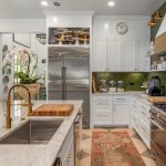 Bright, stylish kitchen with white cabinets, marble countertops, a stainless steel fridge, large gas stove, green herringbone backsplash, brass accents, open shelving, and a colorful rug on the floor.