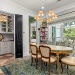 A bright dining area with a round table and four ornate chairs, a blue patterned rug, a bookshelf, and a window overlooking greenery. Double black doors reveal a small nook with shelves and cabinets.