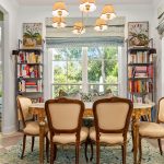 Elegant dining room with six vintage chairs around a round wooden table, bookshelves on either side, large windows letting in natural light, a blue rug, and a modern chandelier overhead.