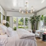 A bright, elegant bedroom with a large bed covered in white linens, two armchairs and a side table by tall windows, green accents in the decor, a modern chandelier, and leafy indoor plants.