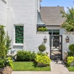 A charming white brick house with green plants, shrubs, and small trees along the walkway, decorative lanterns on the wall, a black gate, and two stone dog statues by the entrance.