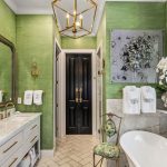 Elegant bathroom with green walls, marble countertops, a freestanding bathtub, gold fixtures, wall art, large mirrors, a floral chair, fresh white towels, and large windows letting in natural light.