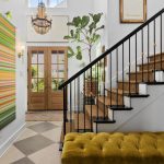 Bright entryway with a large, colorful striped painting, wooden staircase with black railing, green tufted bench, potted plant, and double wooden doors with glass panels. A chandelier hangs from the high ceiling.