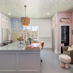 A bright, airy workspace with a large island desk, comfy chairs, art supplies, and colorful artwork on blush pink and white walls. Multiple windows let in natural light, creating a welcoming, creative atmosphere.