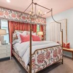 A bedroom with a floral-patterned canopy bed, white bedding, and pink accent pillows. The room features floral curtains, two white nightstands with lamps, and an ensuite bathroom with a vanity visible in the background.