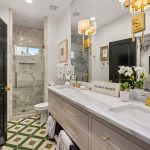 Modern bathroom with double sink vanity, gold fixtures, a large mirror, white cabinetry, green geometric floor tiles, and a glass-enclosed shower. Towels and a potted orchid decorate the counter. Black doors and wall lights add contrast.