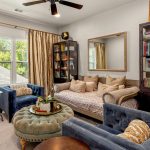 A cozy sitting room with gold curtains, a daybed with many beige pillows, two blue armchairs, a green tufted ottoman, two dark bookcases filled with books, a mirror, and a line drawing artwork on the wall.