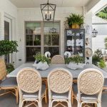 A covered patio with a white dining table and eight wicker chairs. The table is decorated with plants, and a cabinet with towels is against the white brick wall. Greenery surrounds the space, and a pool is visible outside.
