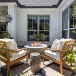 Two cushioned wooden chairs face a small round fire pit table on a stone patio, surrounded by large potted plants, with a white brick house and glass windows in the background.