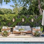 A backyard patio features wicker furniture with white cushions, a round coffee table with flowers, an upright umbrella, and hanging lanterns on a wooden fence covered in climbing greenery, next to a swimming pool.