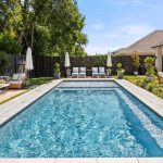 A backyard with a rectangular swimming pool, lounge chairs and umbrellas at the far end, and outdoor seating areas with sofas and tables on the left, surrounded by greenery and a tall wooden fence.