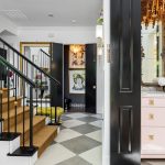 A stylish entryway with black-and-white checkered floors, a staircase with tan carpet and black railings, gold-framed artwork, a bathroom with a pink vanity, and a chandelier. Bright, elegant, and eclectic decor.
