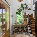 A bright entryway with wooden double doors, a vintage rug, a table with plants and decor, patterned stools, a large beaded chandelier, a green stained glass window, and stairs with black railings.