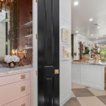 A stylish interior shows a pink vanity with a mirror and ornate sconces, next to an open black door leading to a modern kitchen with bar stools, checkered flooring, and bright decor.