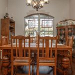 A dining room with a wooden table and six chairs, a chandelier above, a piano on the left, a china cabinet on the right, and stained glass windows letting in daylight. Decor includes framed art and plate displays.